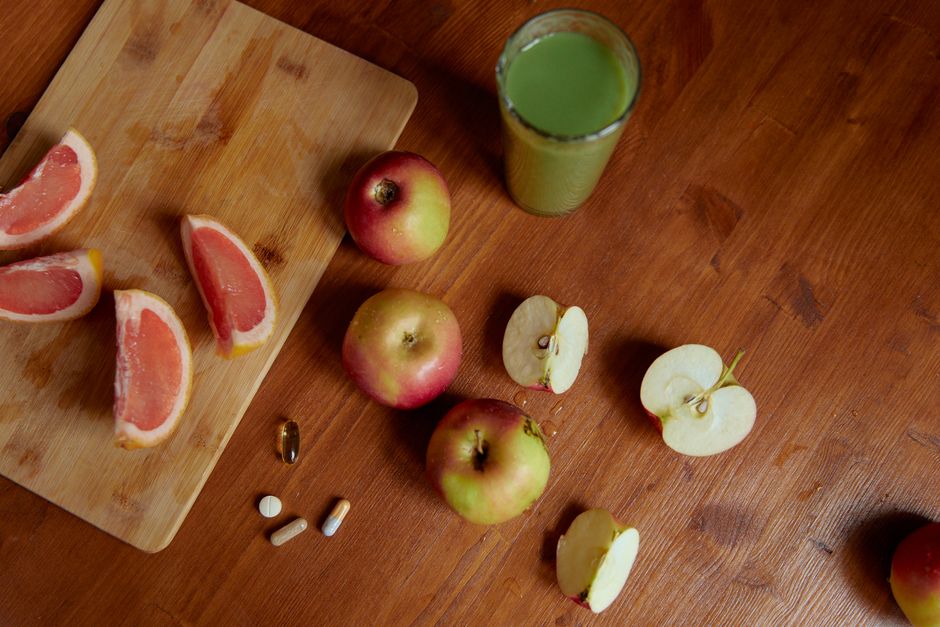 Top view of sliced grapefruit, apples, pills, and a green drink on a wooden table, emphasizing health.