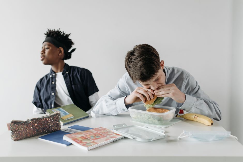 Two teenagers at a school desk, one eating lunch while the other reads. Educational atmosphere.