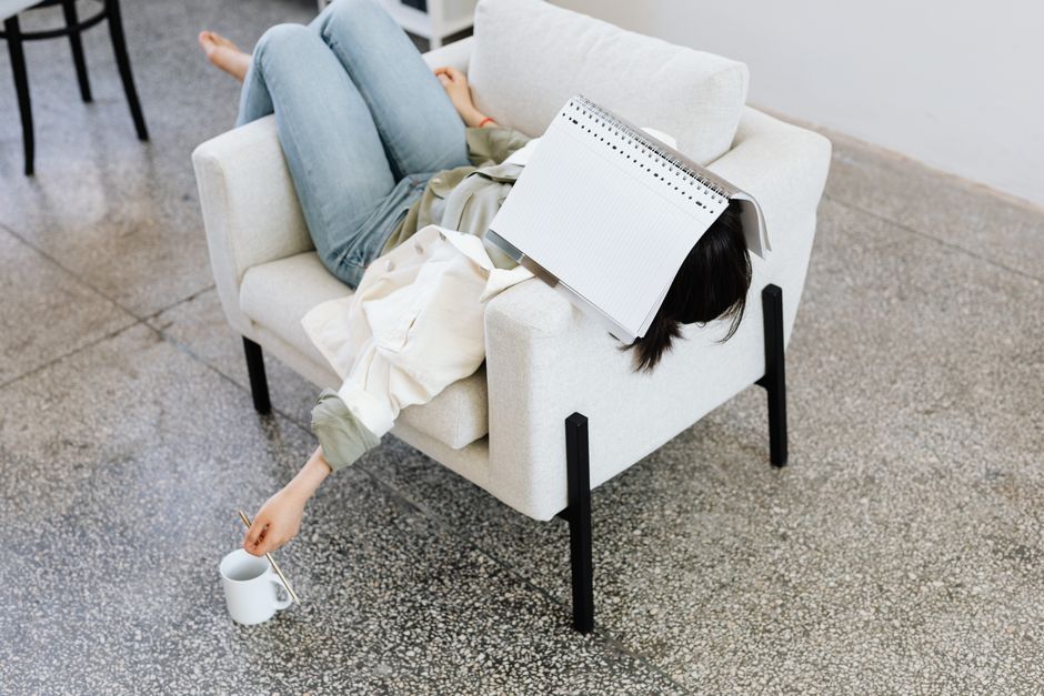 Woman lying on an armchair with a book on face, holding a coffee. Casual and relaxing atmosphere.