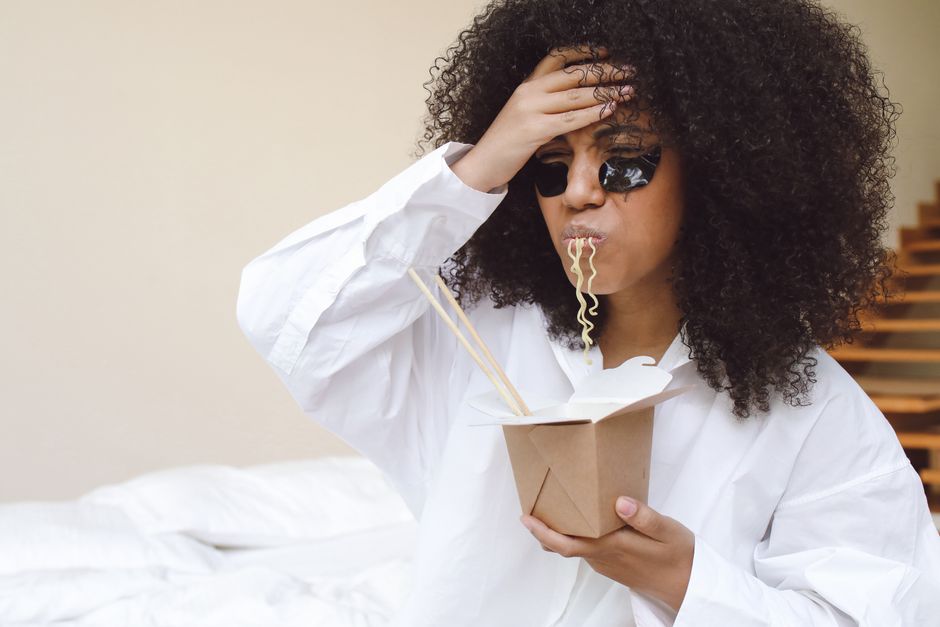 Woman with afro hair eating noodles with under eye patches in a relaxed setting.